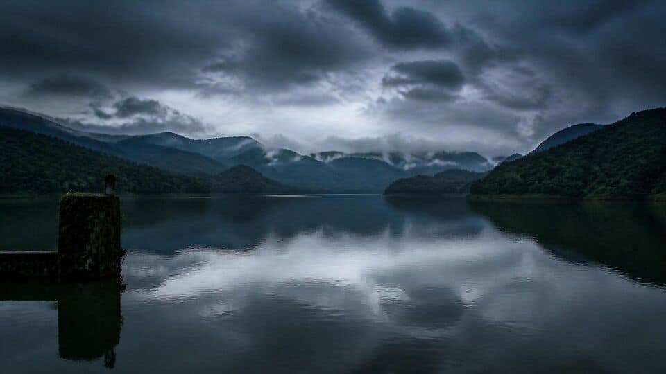 A wide shot of a still lake surrounded by dark, forested mountains under a heavy, grey, cloudy sky, with a small concrete structure visible in the foreground on the left.